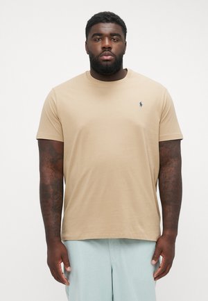 Tan kortærmet t-shirt i blødt stof med rund hals og lille blåt logo på venstre bryst, iført med lyseblå shorts.