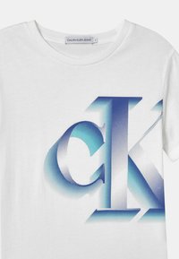 Biała koszulka z okrągłym dekoltem, z dużym niebieskim i turkusowym gradientowym logo "CK" nadrukowanym z przodu oraz metką Calvin Klein Jeans wewnątrz kołnierza.