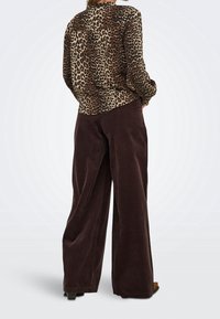 Personne portant une chemise à manches longues imprimée léopard et un pantalon large marron foncé, debout les mains dans les poches, devant un fond uni.