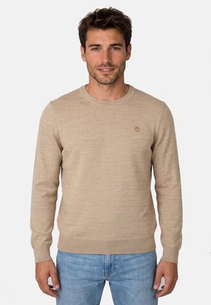Hombre con un suéter beige de cuello redondo y manga larga y jeans azul claro, de pie frente a un fondo claro y liso.