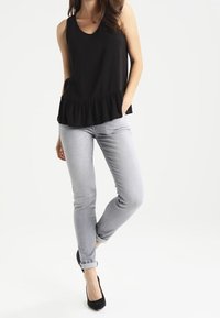 Mujer con una camiseta sin mangas negra con volantes en el dobladillo, jeans ajustados gris claro arremangados y zapatos de tacón negro, de pie con una pierna cruzada.