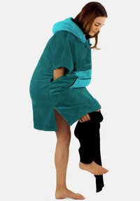 Teal Kapuzentuch aus weichem, saugfähigem Material mit Seitenschlitzen. Eine Person zieht schwarze Hosen an einem Bein an und zeigt eine barfüßige Haltung.