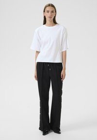 Top alb cu mânecă scurtă, cu o croială relaxată, asociat cu pantaloni negri cu butoane laterale; ambele au texturi netede și un design minimal.