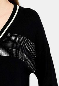 Maglione nero con scollo a V; presenta una fascia testurizzata di borchie lucide sul petto e righe bianche di accento sul colletto.