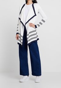 Biały kardigan z granatowymi akcentami i paskowanymi detalami, zestawiony z szerokimi ciemnoniebieskimi dżinsami i białymi butami. Długie rękawy i otwarty fason.