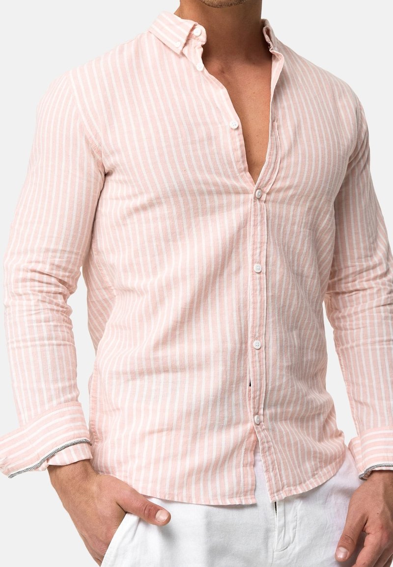 Mann trägt ein hellrosa und weiß gestreiftes Langarmhemd mit teilweise geknöpftem Kragen und hat die Hände in den Taschen von weißen Hosen.