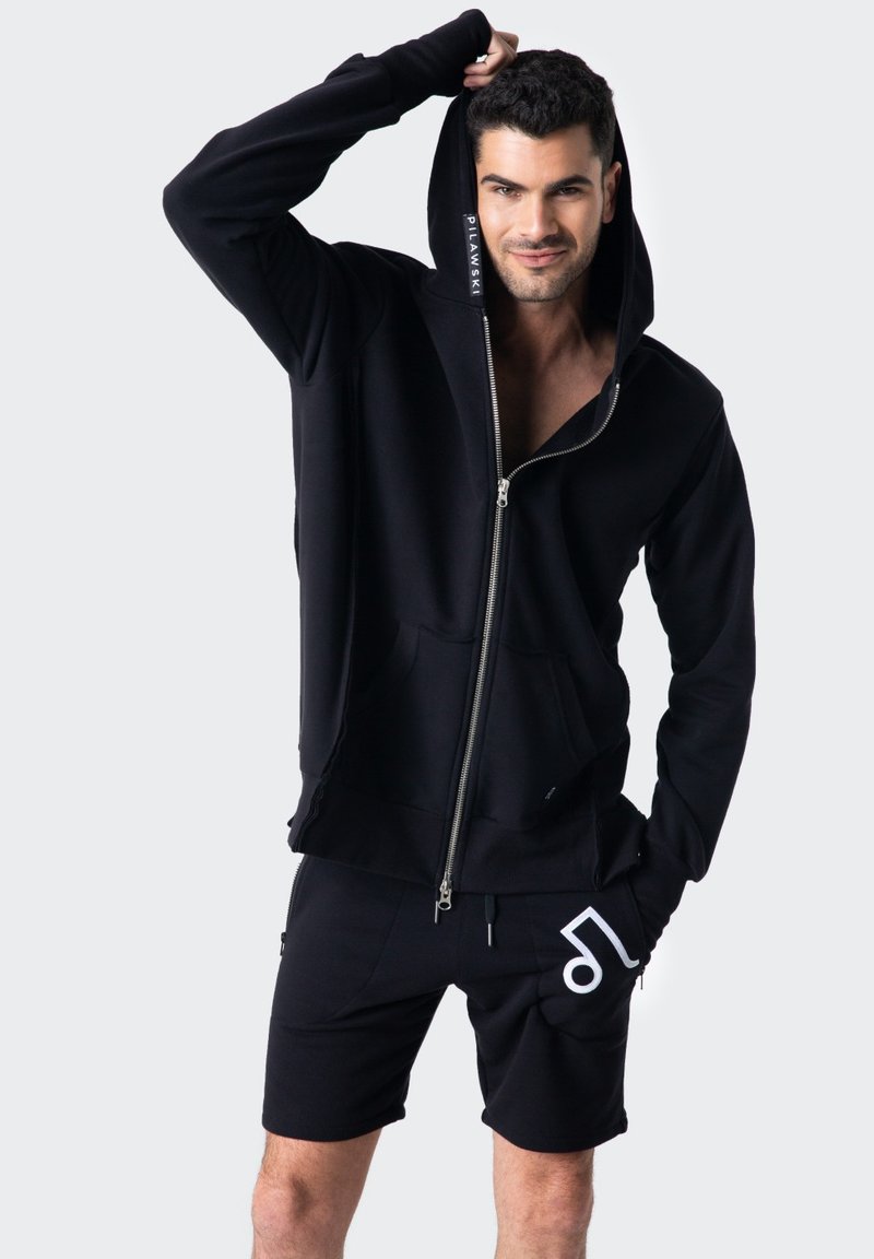Czarna bluza z kapturem na zamek, z kieszeniami z przodu i logo na rękawie, w połączeniu z czarnymi szortami z białym wzorem graficznym.