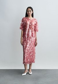 Femme portant un haut rose semi-transparent à sequins et une jupe midi assortie, associée à des escarpins blancs pointus, debout contre un mur uni.