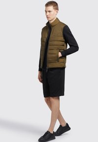 Maslinasto zeleni puffer prsluk s visokim kragnom, zatvaranjem na zip i dva bočna džepa, nošen preko crne majice dugih rukava i crnih šortseva.