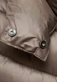 Primer plano de una chaqueta de plumas marrón, mostrando su textura suavemente acolchada y dos botones a presión negros en el dobladillo de la manga.