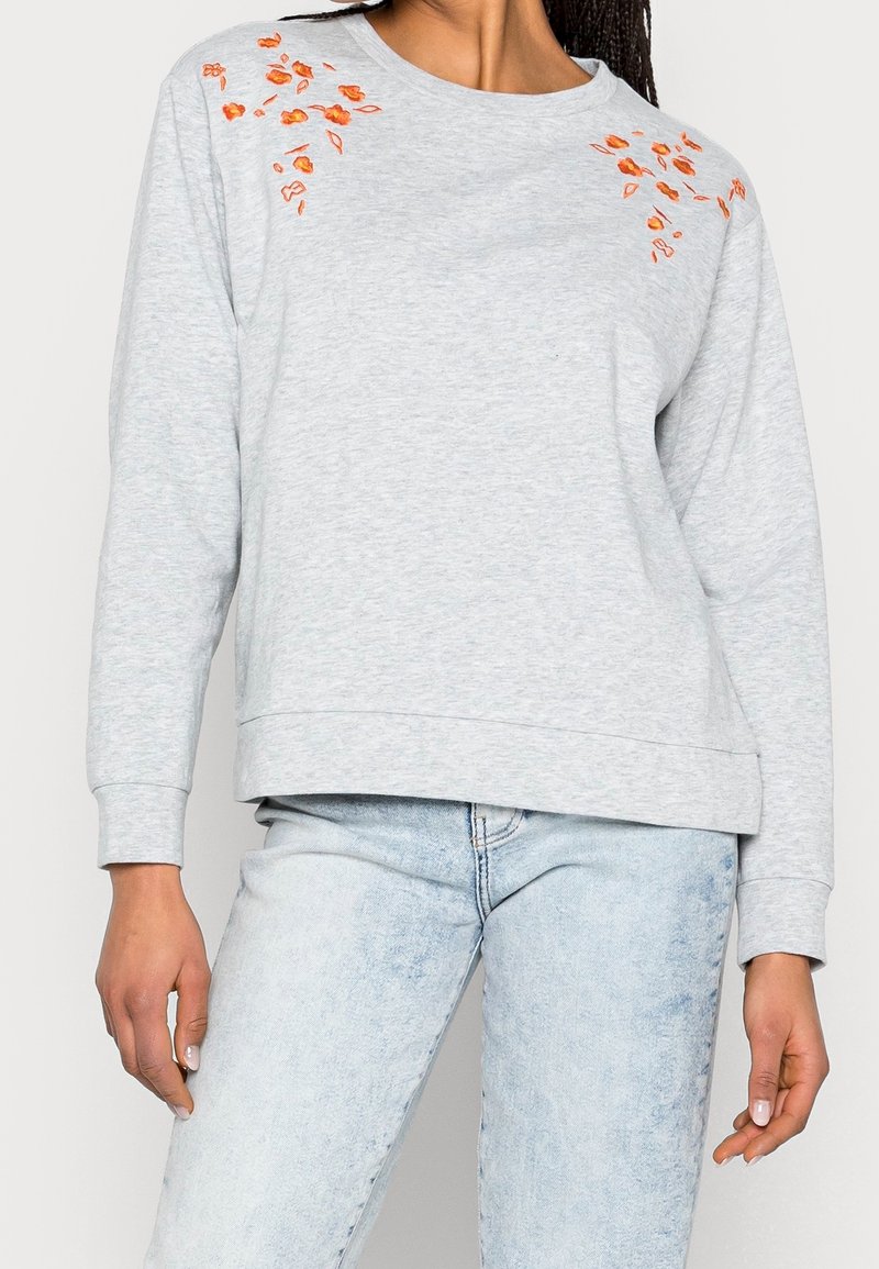 Sweatshirt gris clair à manches longues. Équipé de détails floraux orange brodés sur les épaules. Tissu doux, coupe décontractée.