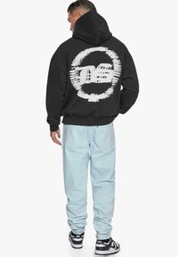 Felpa nera con cappuccio caratterizzata da una grande grafica bianca sul retro, abbinata a jeans azzurri chiari e sneaker nere con dettagli bianchi.