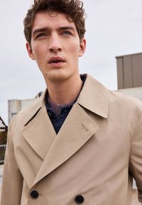 Abrigo trench de doble botonadura beige con grandes botones negros, solapas de muesca y un cuello texturizado, llevado sobre un suéter de punto azul oscuro.