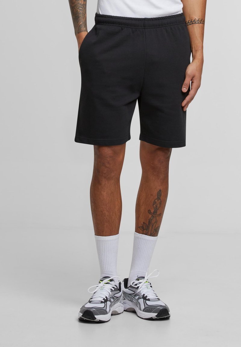 Man wearing black shorts, white crew socks, and gray athletic shoes, standing with hands in pockets against a plain gray background.