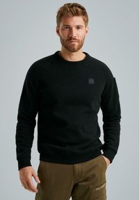 Schwarzer Rundhalsausschnitt-Pullover mit gerippten Bündchen und Saum, mit einem Patch auf der Brust und einer Utility-Tasche am Ärmel. Getragen mit beigefarbenen Cargo-Hosen.
