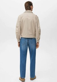 Beige jasje met lange mouwen en een rechte snit, gecombineerd met blauwe spijkerbroeken. De stof lijkt glad met minimale hardware.