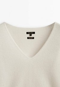 Sweter z kaszmiru w kolorze kremowym z dekoltem w kształcie V, z prążkowanym kołnierzem oraz czarnymi metkami marki "Massimo Dutti" i "100% Kaszmir Wyprodukowano w Chinach."