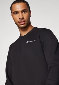 Sweatshirt noir en mélange de coton, avec un col rond et un logo "Champion" brodé en blanc et rouge sur la poitrine.