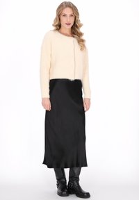 Kobieta z luźnymi falującymi blond włosami, ubrana w kremowy kardigan na guziki, czarną satynową spódnicę midi i czarne skórzane botki, stojąca na białym tle.