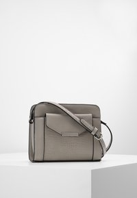 Szara skórzana torba na ramię z przednią kieszenią o fakturze oraz regulowanym paskiem, prezentowana na białym podium.