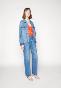 Casaco de denim com lavagem clara, por cima de uma blusa de bolinhas vermelhas, combinado com jeans de perna reta de cintura alta e sapatos de salto branco com tiras.
