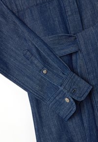 Denim-Bekleidungsstück in tiefblauem Farbton, mit Knöpfen am Ärmel, einem gebundenen Taillenbereich und einem strukturierten Stoff, der horizontale Webung zeigt.
