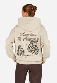 Μπεζ φούτερ με μεγάλη, γραφική εκτύπωση δύο πεταλούδων και το κείμενο "Μείνε πιστός στον εαυτό σου" στην πλάτη. Μαλακό, υφασματώδες υλικό.