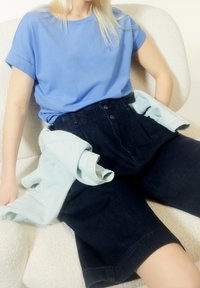 Jasnoniebieski t-shirt z krótkim rękawem w połączeniu z ciemnymi, wysokim stanem, jeansowymi szortami z zapięciem na guziki z przodu oraz jasnoniebieską kurtką ułożoną na nogach.