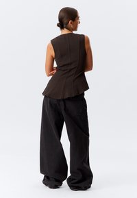 Brun overdelsbluse uten ermer med teksturert design, tettsittende liv, og utsvingt hem, kombinert med løse svarte høytlivede jeans med baklommer.
