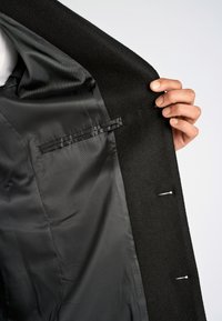 Abrigo de lana negro con un suave forro de satén negro, que muestra bolsillos interiores y detalles de costura, sostenido abierto por una mano.