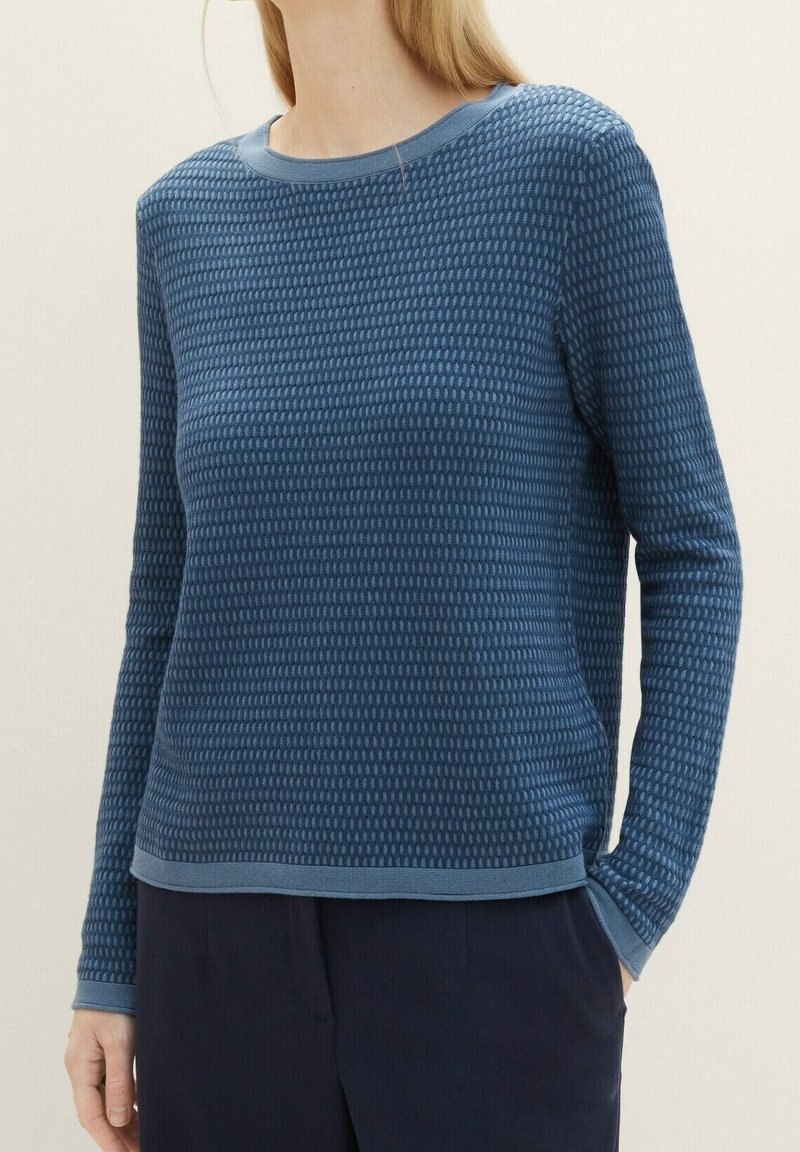Femme portant un pull bleu texturé ajusté à manches longues avec un col rond et un pantalon sombre, debout avec une main dans la poche.