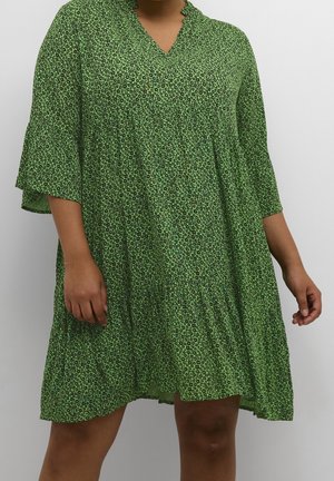Femme portant une robe verte ample longueur genou avec de petits motifs floraux noirs et des manches papillon trois-quarts sur un fond uni.