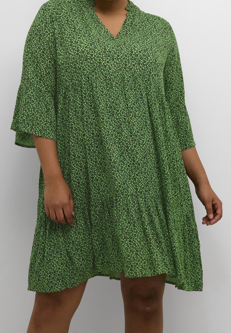 Femme portant une robe verte ample longueur genou avec de petits motifs floraux noirs et des manches papillon trois-quarts sur un fond uni.