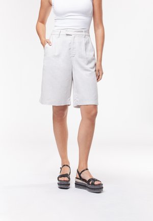 Donna che indossa pantaloncini bianchi a vita alta fino al ginocchio con tasche, una canotta bianca senza maniche e sandali neri con plateau e cinturini alla caviglia.