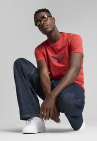 Rød kortærmet t-shirt med mørk skrift og logo, parret med løse mørkeblå jeans og hvide sneakers. Personen bærer gule solbriller.
