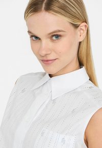 Blusa senza maniche bianca con colletto a bottoni, caratterizzata da linee verticali di accenti argentati e un leggero lucido sul tessuto.