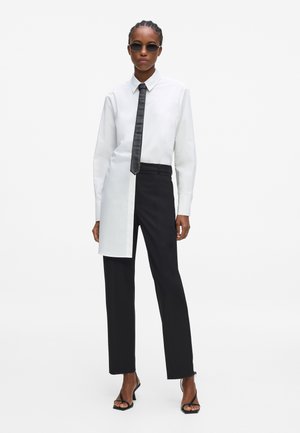 Donna che indossa una camicia bicolore nero e bianco, cravatta nera, pantaloni neri, sandali neri con tacco e occhiali da sole, in piedi su sfondo bianco.