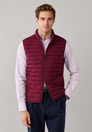 Man draagt een bordeauxkleurig gewatteerd vest over een lichtroze gestreept overhemd met knopen en marineblauwe broek tegen een effen achtergrond.
