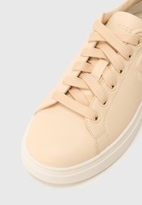 Μπεζ δερμάτινο αθλητικό παπούτσι με στρογγυλεμένη μύτη, επίπεδα κορδόνια και μεταλλικά ρούχιστρα. Διαθέτει υφασμένη επιφάνεια και λευκή ρηχτή σόλα.