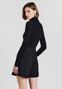 Maglietta nera a maniche lunghe con colletto alto abbinata a pantaloni corti neri a vita alta, con una texture liscia e un design minimalista. Dettagli con anelli in argento.