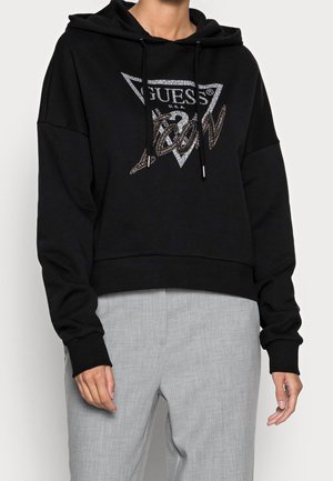 Personne portant un sweat à capuche noir avec un logo "GUESS U.S.A." et des lettres métalliques, associé à un pantalon gris clair.