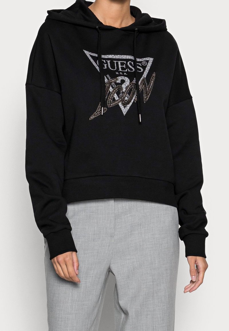 Personne portant un sweat à capuche noir avec un logo "GUESS U.S.A." et des lettres métalliques, associé à un pantalon gris clair.