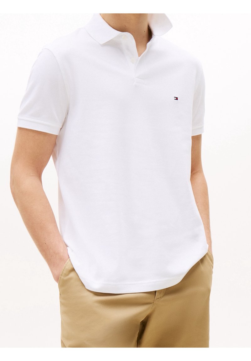 T-shirt polo blanc avec un col structuré, deux boutons et un petit logo sur la poitrine gauche. Porté avec un pantalon khaki, mettant en valeur un style décontracté.