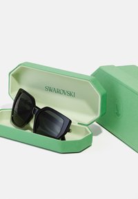 Suured, nurksed musta raami ja tumedate läätsedega prillid, mis on paigutatud rohelisse tekstuuriga säilituskarpi, mille sees on graveeritud "SWAROVSKI".