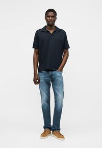 Mann står iført en mørk marineblå kortermet skjorte, blå jeans og lysebrune joggesko med én hånd i lommen.