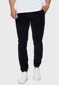 Pantalons jogger en velours côtelé noir avec une taille élastique et des poignets élastiques, présentant une finition texturée et des poches latérales, portés avec des baskets blanches.