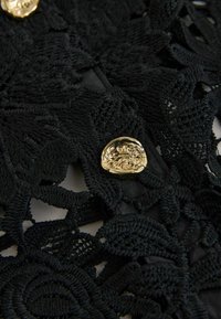 Sort blonderstof med indviklede blomster-mønstre, der har en fremtrædende guldknap som lukningsdetalje. Tekstureret overflade med lagdelt design.