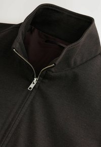 Svart zip-up jacka med hög krage, slätt tyg och en silverdragkedja. Inre foder är mörklila. Minimalistisk design utan mönster.