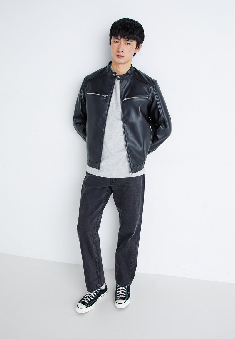 Schwarze Lederjacke mit Reißverschlusstaschen, graues T-Shirt, dunkle Jeans und schwarze Sneaker mit weißen Sohlen, vor einem weißen Hintergrund stehend.