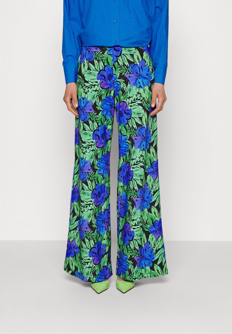 Pantalon large avec un motif floral bleu vif et violet et des feuilles vertes, porté avec une chemise bleue et des chaussures pointues vert clair.